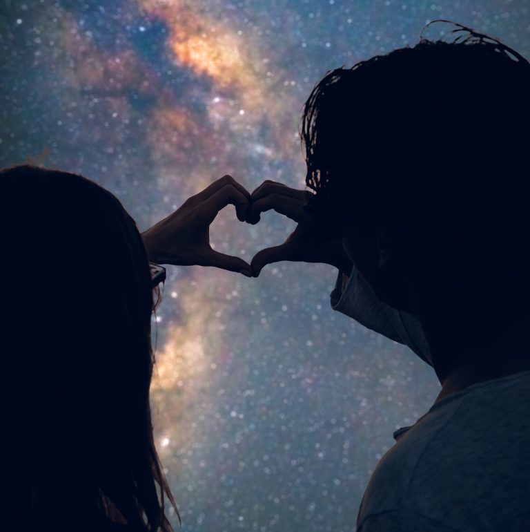 man and woman form a heart with their hands over Milky Way