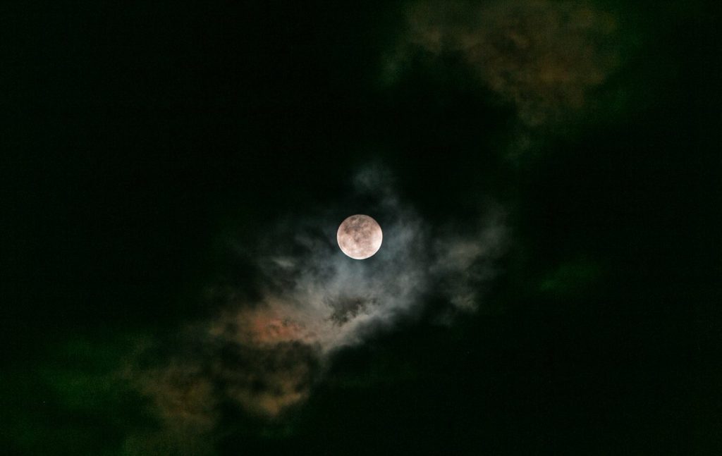 Full Moon in a dark sky viewed through the clouds. The clouds appear reddish next to the moon